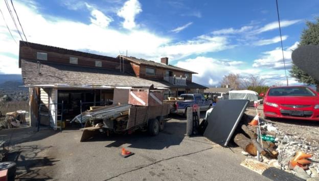 A photo of a weathered two story home on a Kelowna street with piles of building materials and a truck in front loaded with co<em></em>nstruction equipment. 