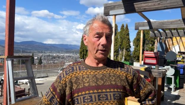 A 73-year-old man stands in his back yard on a cloudy day with hills in the distance. He is standing in a cluttered yard with a lot of co<em></em>nstruction material around him. 